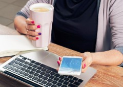 Obsession A woman sitting in front of a laptop, looking at a smartphone in one hand while holding a coffee in the other